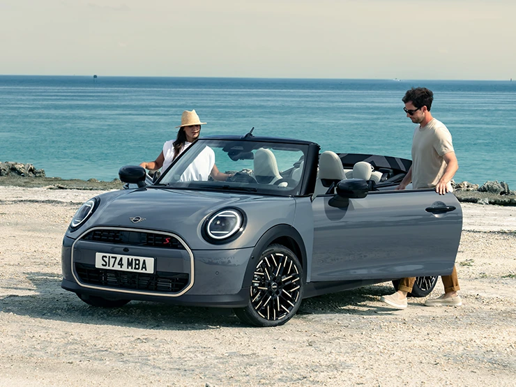 MINI Cooper Cabrio i Copper Grey på en strand med to personer ved siden av, sett forfra fra venstre side.