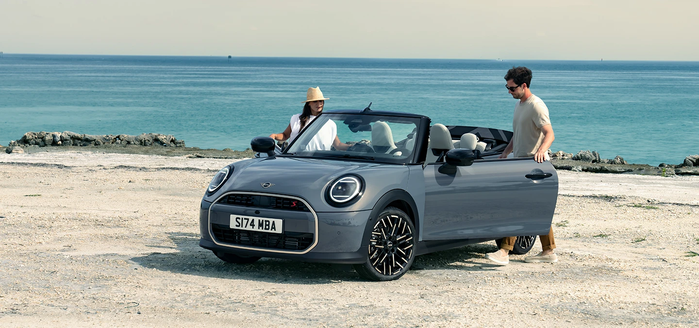 MINI Cooper Cabrio i Copper Grey på en strand med to personer ved siden av, sett forfra fra venstre side.