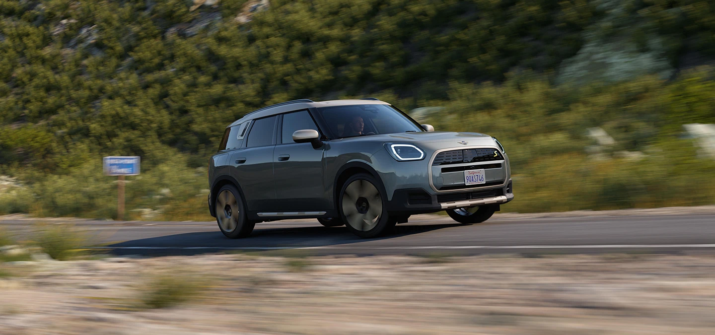 En terrengkjørende helelektrisk MINI Countryman i Smokey Green med tak i Vibrant Silver, sett fra siden.