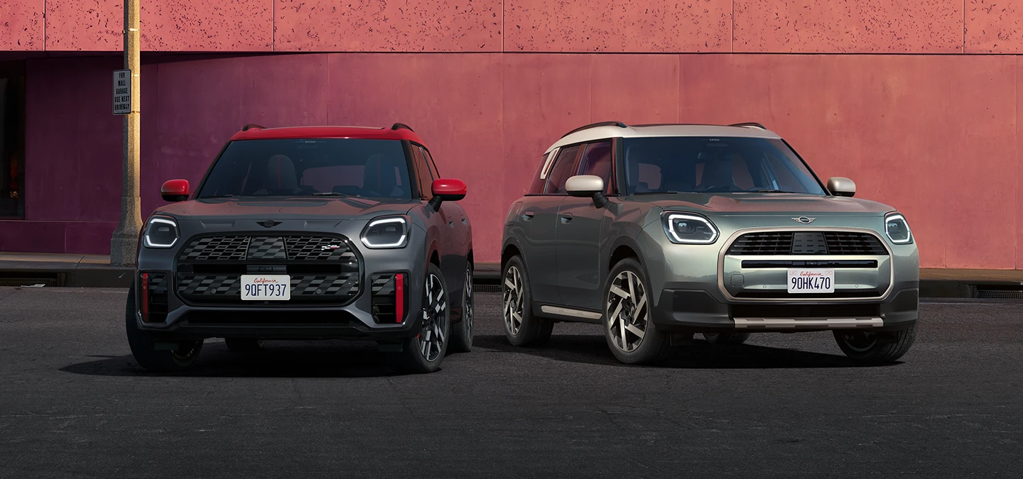 MINI Countryman JCW i Legend Grey med rødt tak og MINI Countryman i Smokey Green med tak i Vibrant Silver som står ved siden av hverandre, sett forfra.