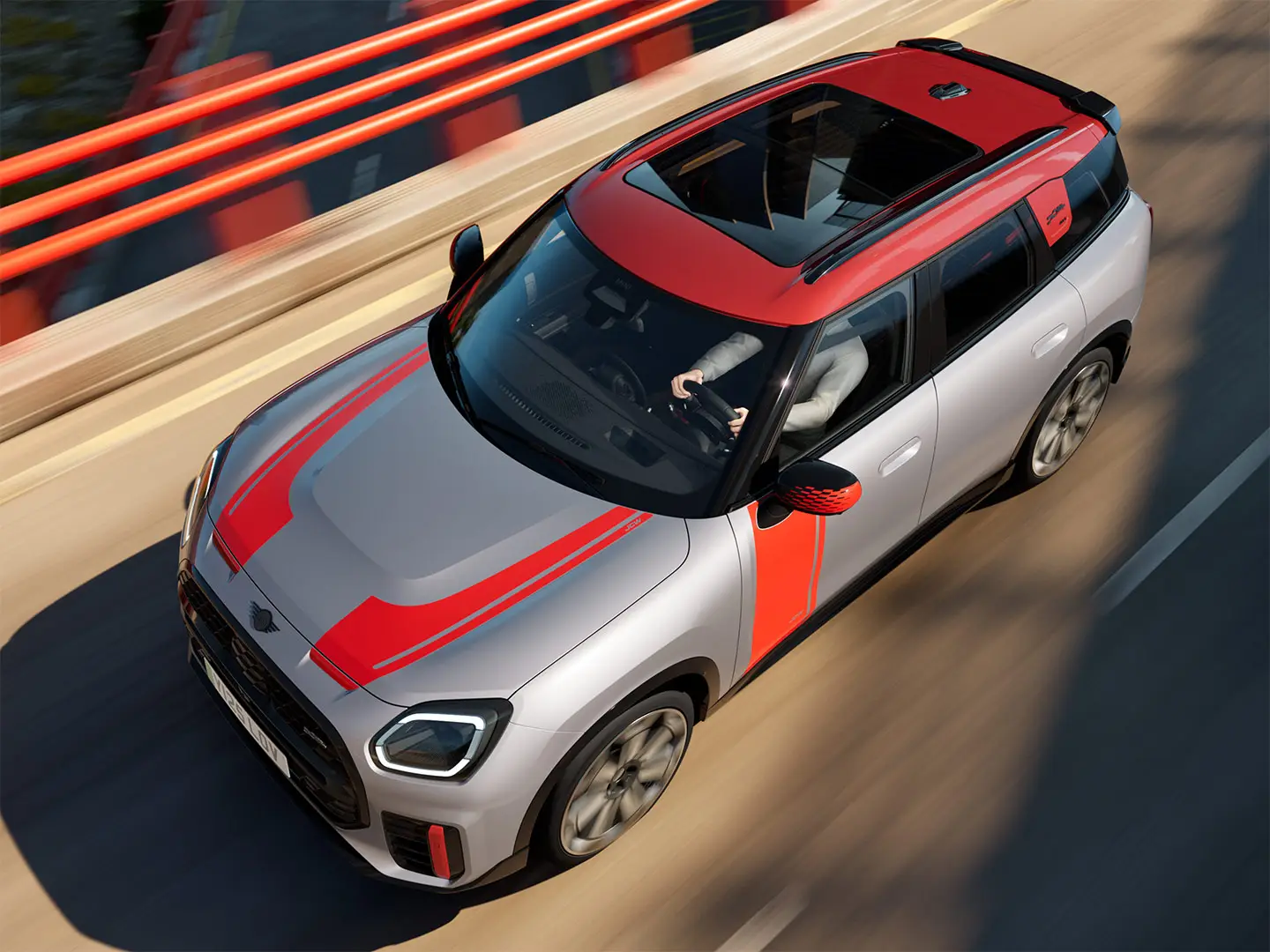 MINI Countryman John Cooper Works i Melting Silver med sidespeildeksel, klistremerker på siden og på panseret, tak og vinge i Chili Red – sett fra siden forfra mens den kjører i en bygate.