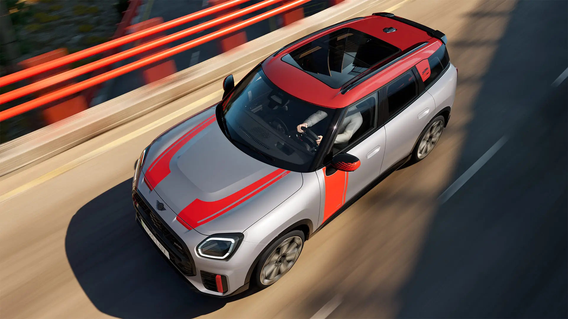 MINI Countryman John Cooper Works i Melting Silver med sidespeildeksel, klistremerker på siden og på panseret, tak og vinge i Chili Red – sett fra siden forfra mens den kjører i en bygate.