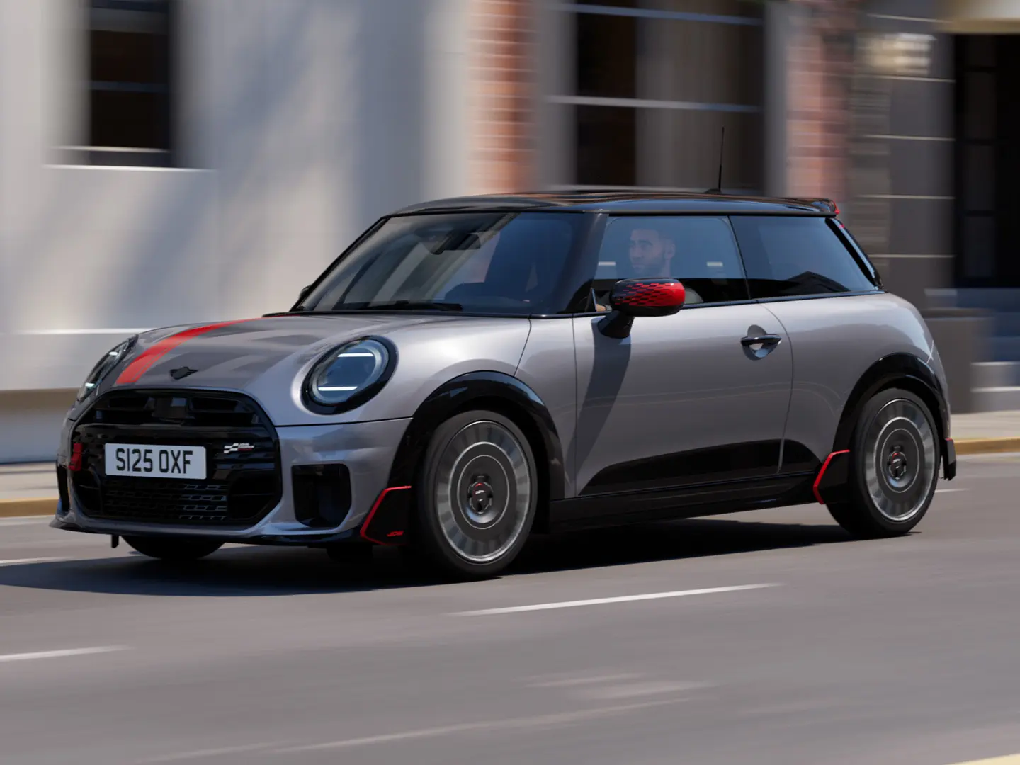 3-dørs MINI Cooper i Melting Silver med John Cooper Works sidespeildeksel og panserklistremerke i Chili Red, takspoiler og vinge i svart – sett fra siden forfra mens den kjører i en bygate.
