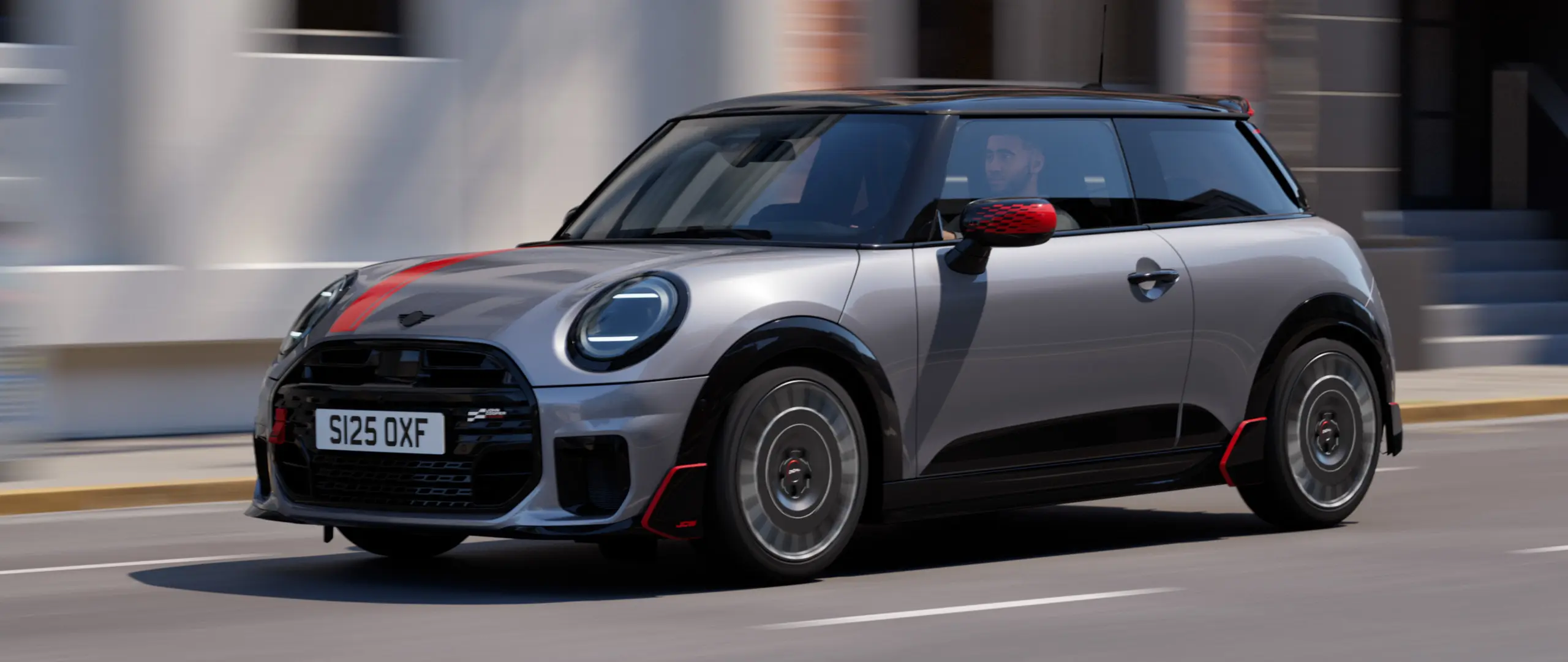 3-dørs MINI Cooper i Melting Silver med John Cooper Works sidespeildeksel og panserklistremerke i Chili Red, takspoiler og vinge i svart – sett fra siden forfra mens den kjører i en bygate.