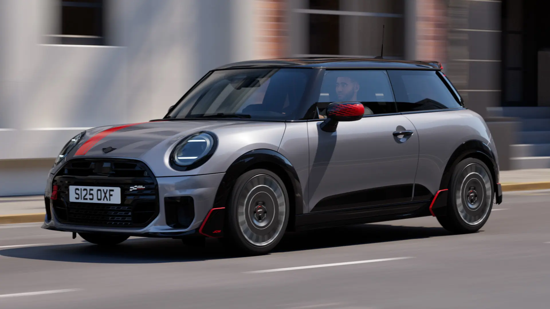 3-dørs MINI Cooper i Melting Silver med John Cooper Works sidespeildeksel og panserklistremerke i Chili Red, takspoiler og vinge i svart – sett fra siden forfra mens den kjører i en bygate.